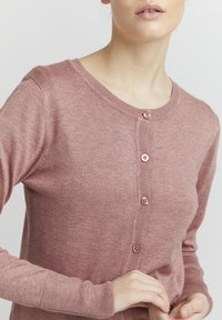 Roza barva pletenega cardigan z okroglim izrezom, sprednjim gumbom in dolgimi rokavi, ki imajo rebraste manšete in robe. Mehka tekstura in prilagojena oblika.