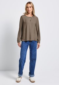 Blusa long-sleeve verde oliva con scollatura a chiave, abbinata a jeans blu a gamba dritta e sneakers bianche con dettagli beige.
