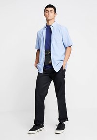 Cămașă bleu cu mâneci scurte peste un tricou bleumarin, combinată cu pantaloni negri și adidași. Include o borsetă cu un model camuflaj.