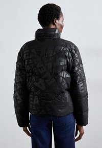 Zwarte pufferjack met een gestructureerd geometrisch patroon, met een hoge kraag en zachte, gewatteerde stof. Draag je over blauwe jeans.
