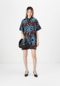 Krótkosleeve, oversize sukienka w kolorze brązowym z niebieskim wzorem promieni słonecznych. Posiada kołnierzyk i zapięcie na guziki. Połączona z czarną torebką i obcasami.