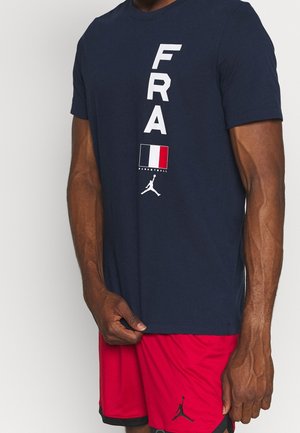 Man wearing a navy blue shirt with vertical "FRA" text, French flag, and basketball logo, paired with red shorts featuring a black basketball logo.