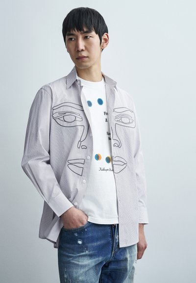 Chemise légère à rayures avec un motif de contour de visage, portée sur un t-shirt blanc graphique avec des points bleus et orange. Portée avec un jean déchiré.