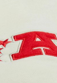 Der cremefarbene Stoff zeigt prominent gestickten roten Buchstaben "A" mit weißen Akzenten und umgebenden Sternen. Weiche Textur.