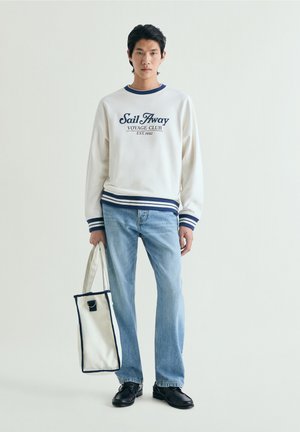 Jeune homme portant un sweat-shirt blanc avec le texte « Sail Away », jean bleu clair, chaussures noires, tenant un sac fourre-tout blanc avec des bordures foncées.