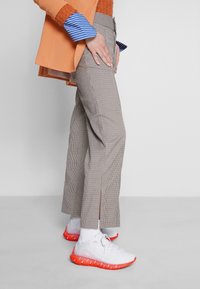 Pantalon à motif pied-de-poule beige et marron, associé à un blazer orange vif. Baskets blanches avec des accents rouges et chaussettes blanches.