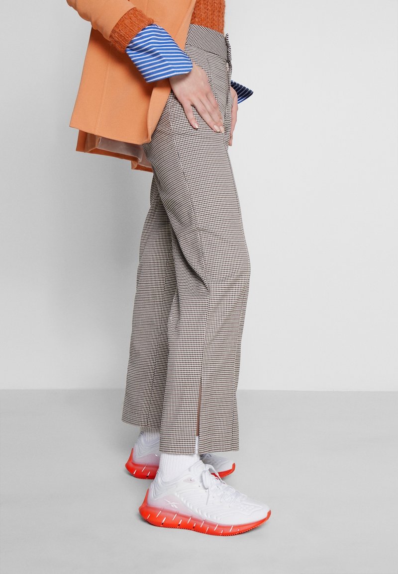 Pantalon à motif pied-de-poule beige et marron, associé à un blazer orange vif. Baskets blanches avec des accents rouges et chaussettes blanches.