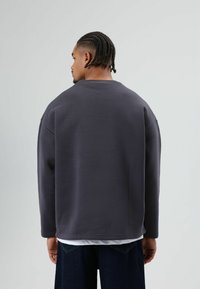 Grijze trui met een ontspannen pasvorm, verlaagde schouders en een ronde halslijn. Glad materiaal, minimale stikseldetails, gedragen over een wit shirt.