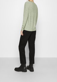 Henkilö, jolla on päällään vaaleanvihreä ribattupinta neule, mustat housut ja mustat paksupohjaiset nilkkasaappaat, seisoo yksinkertaista taustaa vasten.