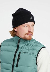 Zielona kamizelka puchowa z zamkiem błyskawicznym z przodu, czarnymi akcentami i logo. Noszona z czarną dzianinową czapką. Powierzchnia ma gładką, izolowaną teksturę.