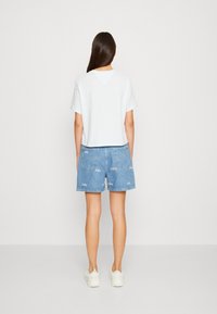 Žena s dlouhými hnědými vlasy, oblečená v bílé košili s krátkým rukávem, modrých denimových šortkách s bílými vzory a bílých teniskách, stojící s otočeným zády k nám.