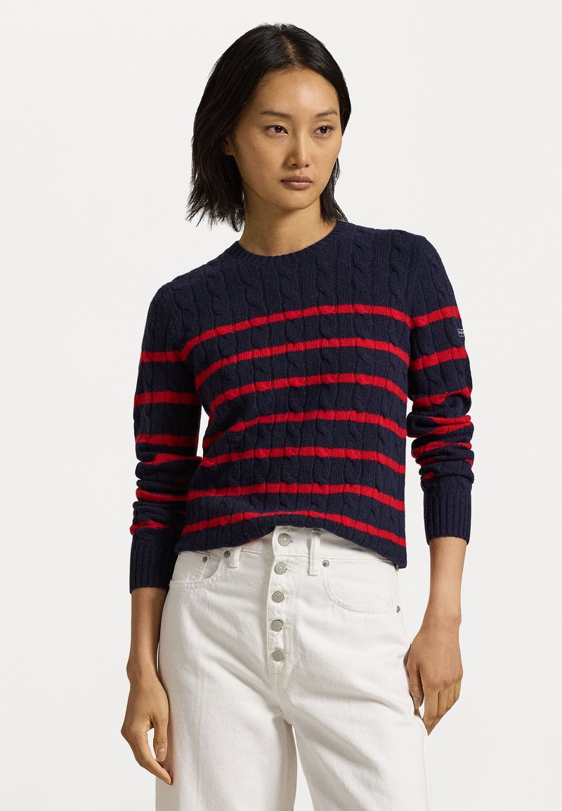 Pull en mailles côtelées bleu marine avec des rayures horizontales rouges, présentant un col rond et des poignets côtelés, associé à un jean taille haute blanc.