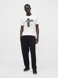 Άνδρας που φοράει λευκό μπλουζάκι Moschino με σχέδιο, μαύρο παντελόνι και μαύρα αθλητικά παπούτσια, στέκεται μπροστά σε απλό φωτεινό φόντο με τα χέρια στις τσέπες.