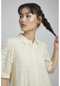 Camicia gialla chiara con dettagli in pizzo, maniche corte a sbuffo e chiusura con bottoni. Presenta un colletto classico e una vestibilità leggermente ampia.