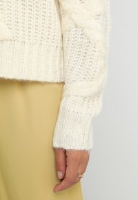 Chunky vit stickad tröja med kabelmönster och ribbade muddarna, som visar upp en mjuk textur. Matchad med ett ljusgult tyg nedanför.