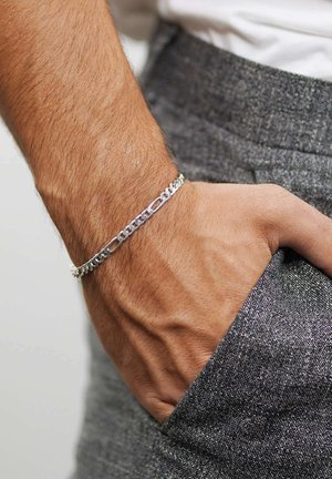 Silberne Gliederkette am Handgelenk, bestehend aus abwechselnden Gliedern mit offenen und soliden Designs. Getragen mit grauen strukturierten Hosen.