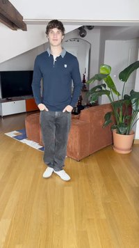 Polo a maniche lunghe blu navy con colletto grigio, jeans grigi e sneaker bianche, in piedi in una stanza con pavimenti in legno, con una pianta e mobili.