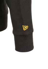 Manica di felpa nera con polsino a coste e piccolo logo giallo ricamato vicino al polso su sfondo bianco.