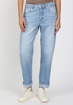 Person trägt hellblaue hochgekrempelte Jeans und weiße Sneaker, steht vor einem einfarbigen hellen Hintergrund.