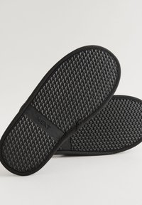 Sandales noires avec une semelle en caoutchouc texturée, comportant un détail logo. Le design est plat et décontracté, avec une surface lisse et des accents minimaux.