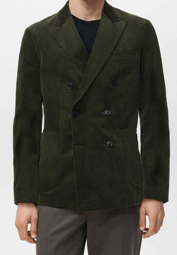 Homme portant un blazer en velours côtelé vert foncé à double boutonnage sur une chemise noire et un pantalon gris, debout avec les bras détendus sur les côtés.
