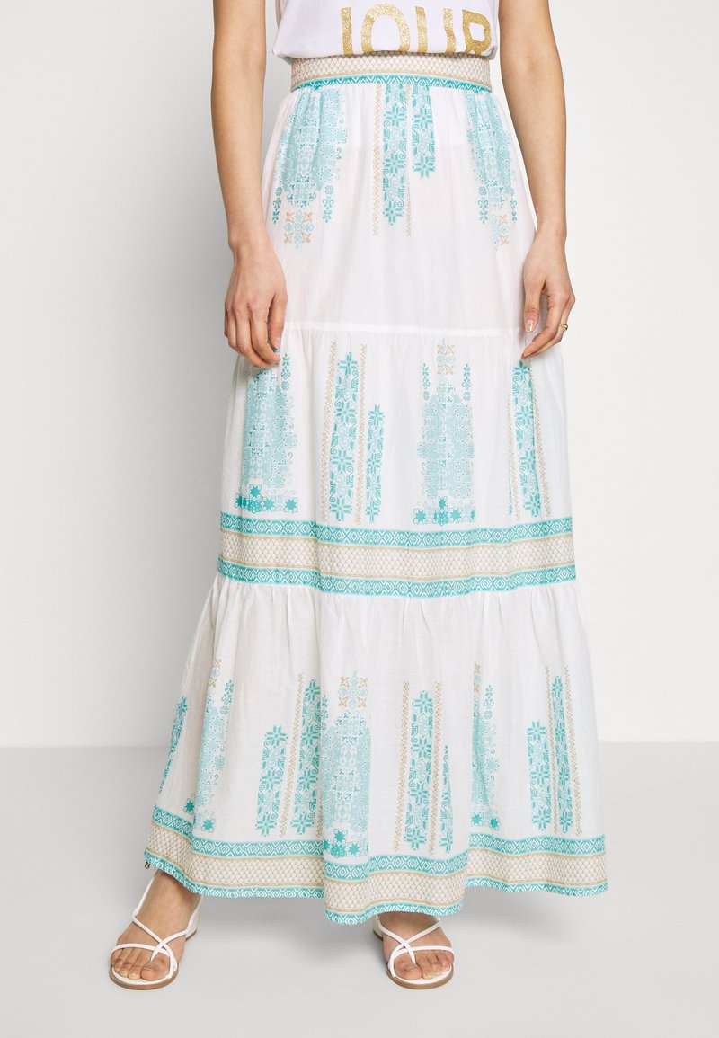 Femme portant une jupe maxi blanche avec des motifs géométriques turquoise et dorés, associée à des sandales blanches à brides et un haut blanc.