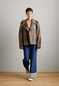 Person som går innendørs iført en oversized brun skinnjakke, oppbrettede vidbukser, hvite sneakers og hoop øredobber foran en ensfarget vegg.