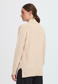 Sieviete ar brūniem matiem astē, tērpusies bēša augstajā kaklā laikā un melnās biksēs, stāv pusprofilā pret vienkāršu gaišu fonu.