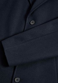 Marinblå ullkappa med en uppslagbar krage, utrustad med en enkel knappstängning och synliga sömmar på ärmslut och fåll. Mjuk textur.