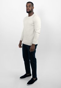 Lichtcrème lange mouwen trui, donkergroene tapered broek en zwarte instapschoenen. Model staat casual tegen een witte achtergrond.
