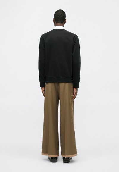 Bărbat văzut de spate, purtând o cămașă neagră cu mâneci lungi, pantaloni bej cu croială largă și pantofi negri pe un fundal alb simplu.