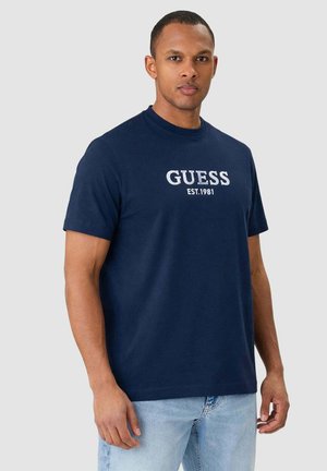 Uomo che indossa una t-shirt Guess blu navy con logo bianco, in piedi davanti a uno sfondo grigio chiaro uniforme.