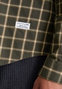 Groen en beige geblokt stof met een wit etiket waarop staat "Marc O' Polo DENIM Gecreëerd met een Scandinavische mindset" op een lange mouw manchet.