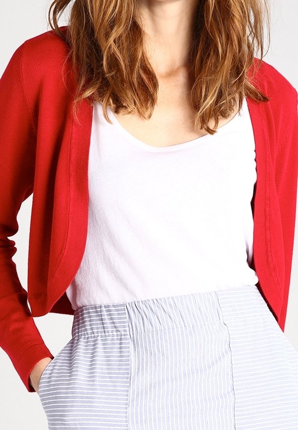 Rød strikket cardigan med sjalskrave, båret over en hvid top. Stribede shorts med høj talje er synlige nedenfor, med elastisk talje.