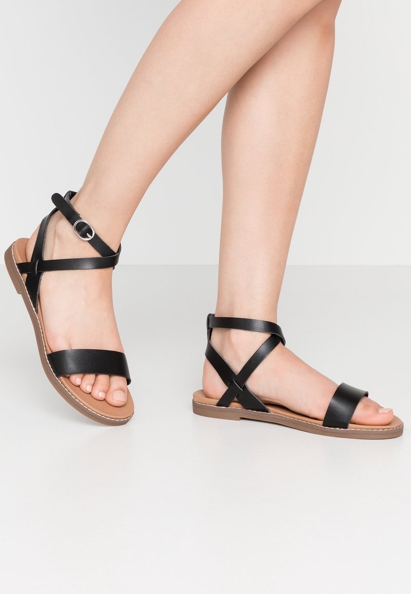 Jalat mengenakan sandal datar kulit hitam dengan tali pergelangan kaki dan gesper, berdiri di atas permukaan berwarna terang.