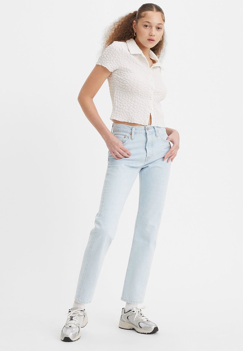 Haut blanc texturé à manches courtes avec col, associé à un jean droit taille haute bleu clair et des baskets en argent.