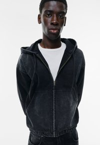 Mustassa denim-hupparissa on vetoketju edessä, kengurupocket ja joustava helma. Sitä on käytetty valkoisen crew neck -t-paidan päällä, jossa on sileä pinta.