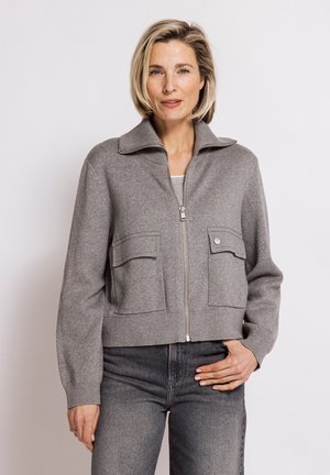 Woman with short blonde hair wearing a grey zip-up jacket with two front pockets and dark grey jeans against a light background.