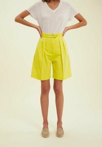Shorts taille haute jaunes vifs avec des plis, maintenus par une ceinture. Texture lisse, design à jambe large. Associés à un t-shirt blanc à col V.