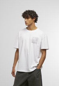 White cotton t-shirt with short sleeves featuring "SPEED DEMON" graphic printed on the left chest. Paired with gray denim shorts.