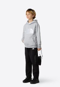 Hanorac gri cu logo alb, pantaloni negri și pantofi negri. Persoana ține o geantă mică neagră cu șnur. Design simplu și casual.