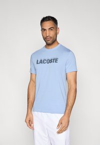 Camiseta de algodón azul claro con mangas cortas y cuello redondo, con un logo negro "LACOSTE" en el pecho. Textura suave.