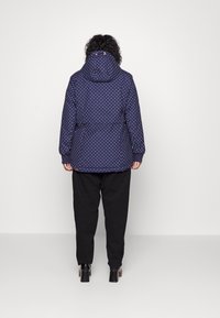 Navy polka dot jacket with a hood, featuring elastic cuffs and a cinched waist for temperature control, styled with black pants and ankle boots.