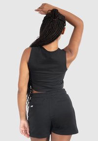 Donna con i lunghi capelli intrecciati, che indossa un top senza maniche nero e pantaloni corti abbinati, in piedi con un braccio alzato dietro la testa.