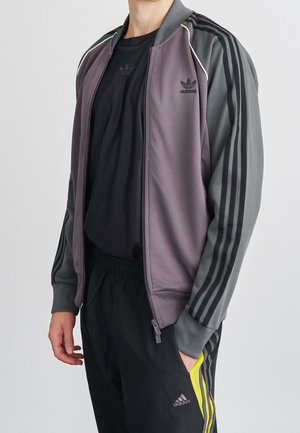 Man draagt een grijs en paars Adidas trainingsjack over een zwart Adidas shirt en zwarte Adidas broek met gele strepen, hand in de zak.