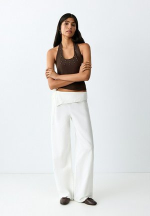 Vrouw met lang donker haar, gekleed in een mouwloze bruine top met studs, witte broek met wijde pijpen en bruine platte schoenen, staand met gekruiste armen.