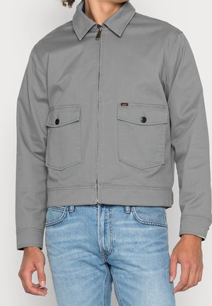 Mann trägt eine graue Reißverschlussjacke mit geknöpften Klappentaschen, kombiniert mit hellblauen Jeans.