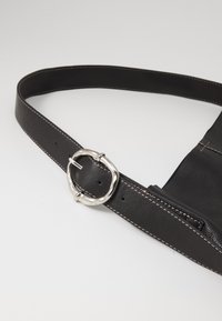 Cinto de couro preto com uma tira larga, apresentando costura em contraste e uma fivela circular prateada. Textura suave com design minimalista.