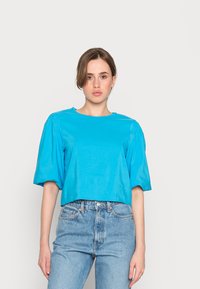 Blusa azul pavilhão curto de cor verde-água com mangas curtas em forma de bola, decote redondo e textura lisa, combinada com jeans claros de cintura alta.