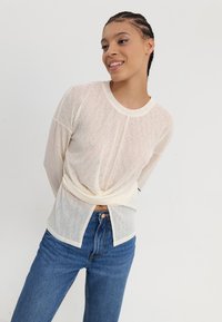 Lichtgewicht, crèmekleurig gebreid topje met lange mouwen en een gedraaide voorzijde, gecombineerd met hooggerimde blauwe jeans.